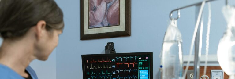 A nurse in blue scrubs examines a medical monitor displaying vital signs in a hospital setting.